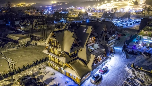 A snow-covered village with traditional chalet-style buildings, a ski resort in the background, and a parking area with cars.