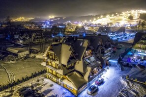 Ein schneebedecktes Dorf mit traditionellen Chalets, ein Skigebiet im Hintergrund und ein Parkplatz mit Autos.