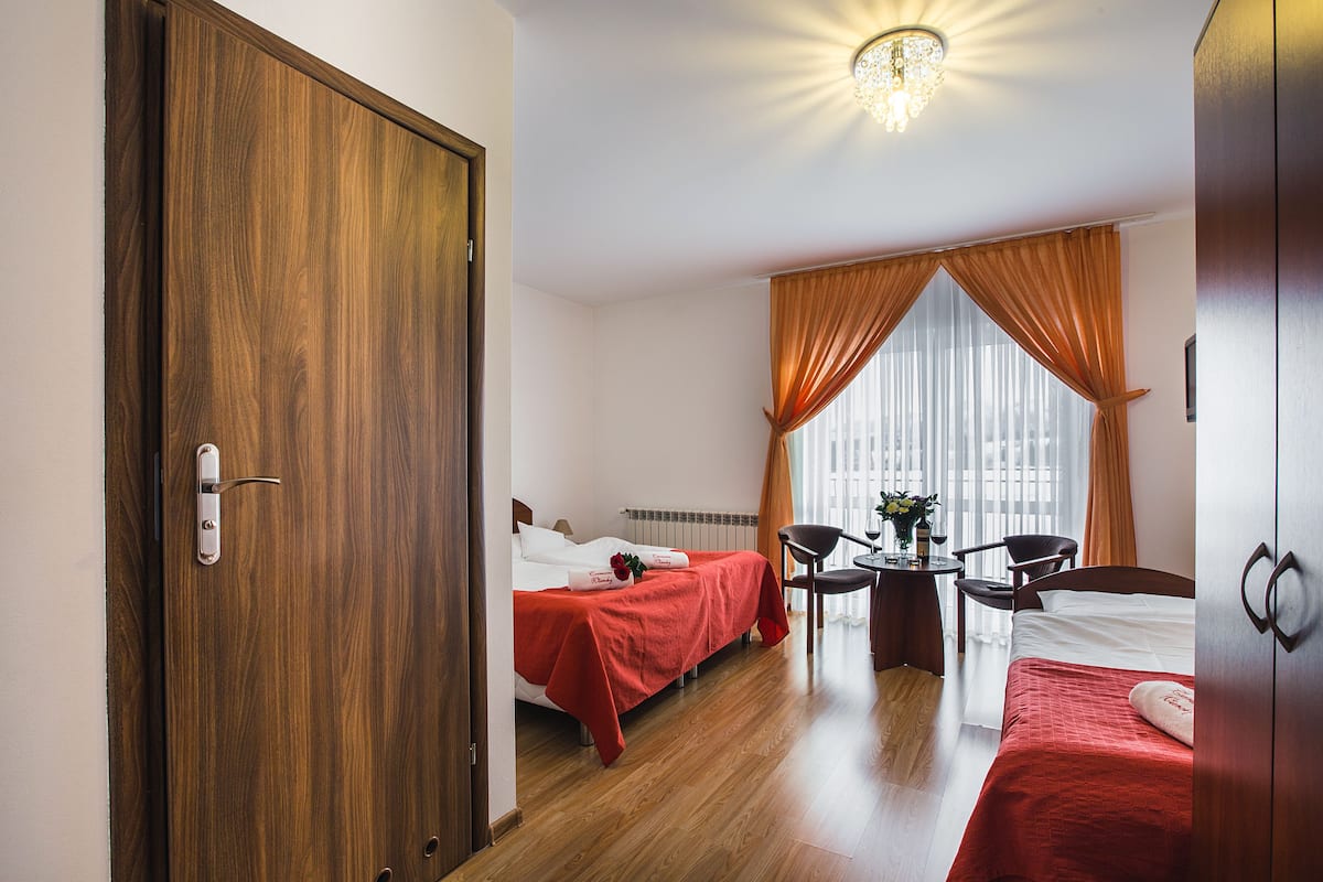Ein Hotelzimmer mit zwei Betten, einer Holztür, einer Kristallleuchtre, einem kleinen Tisch mit einer Vase und einem Fenster mit Vorhängen.