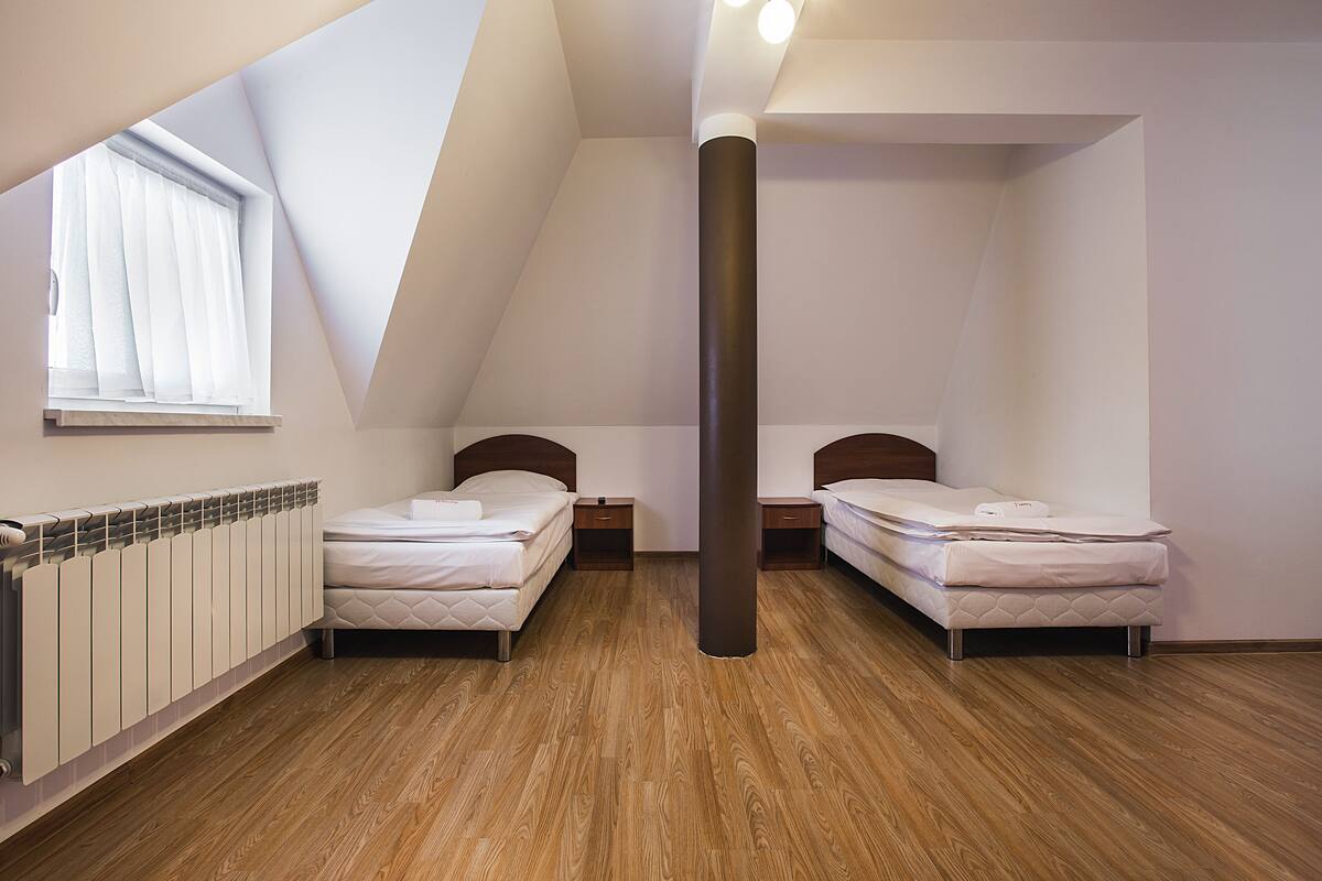 Ein Zimmer mit zwei Betten, einem Fenster mit transparenten Vorhängen, einem Heizkörper und Holzboden.