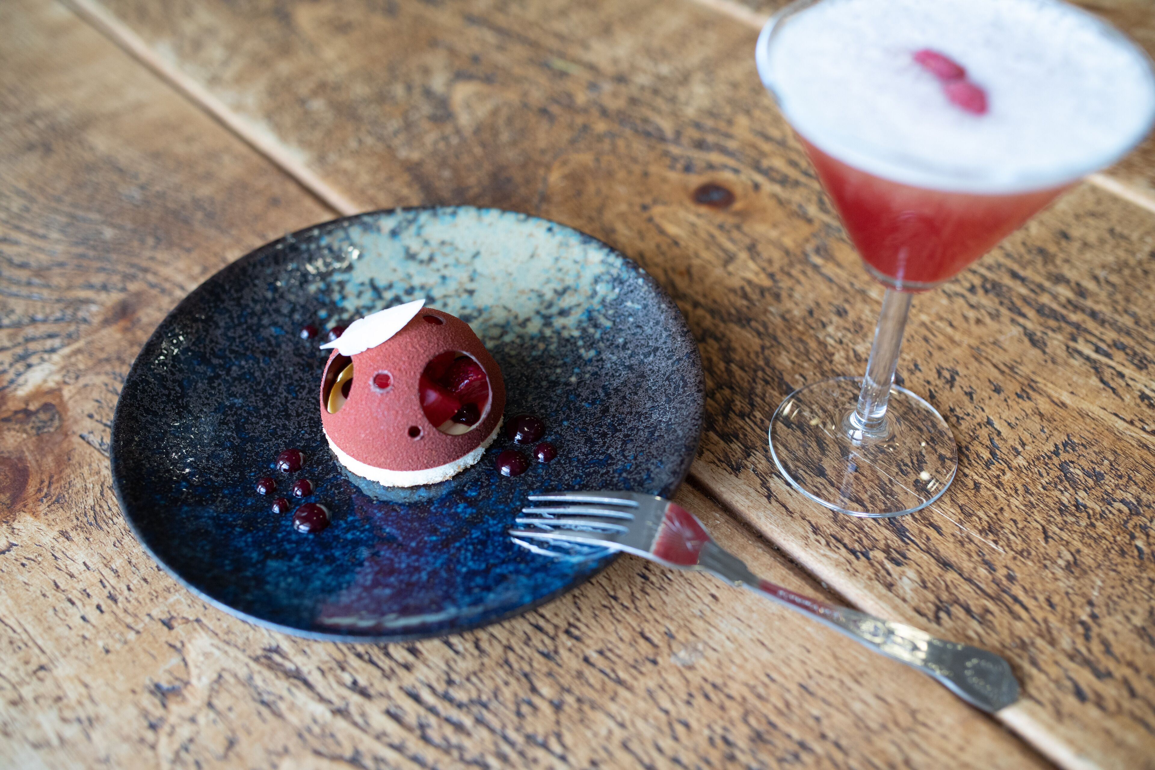 A dessert on a blue plate with a fork beside it, and a martini glass with a cherry on the rim.