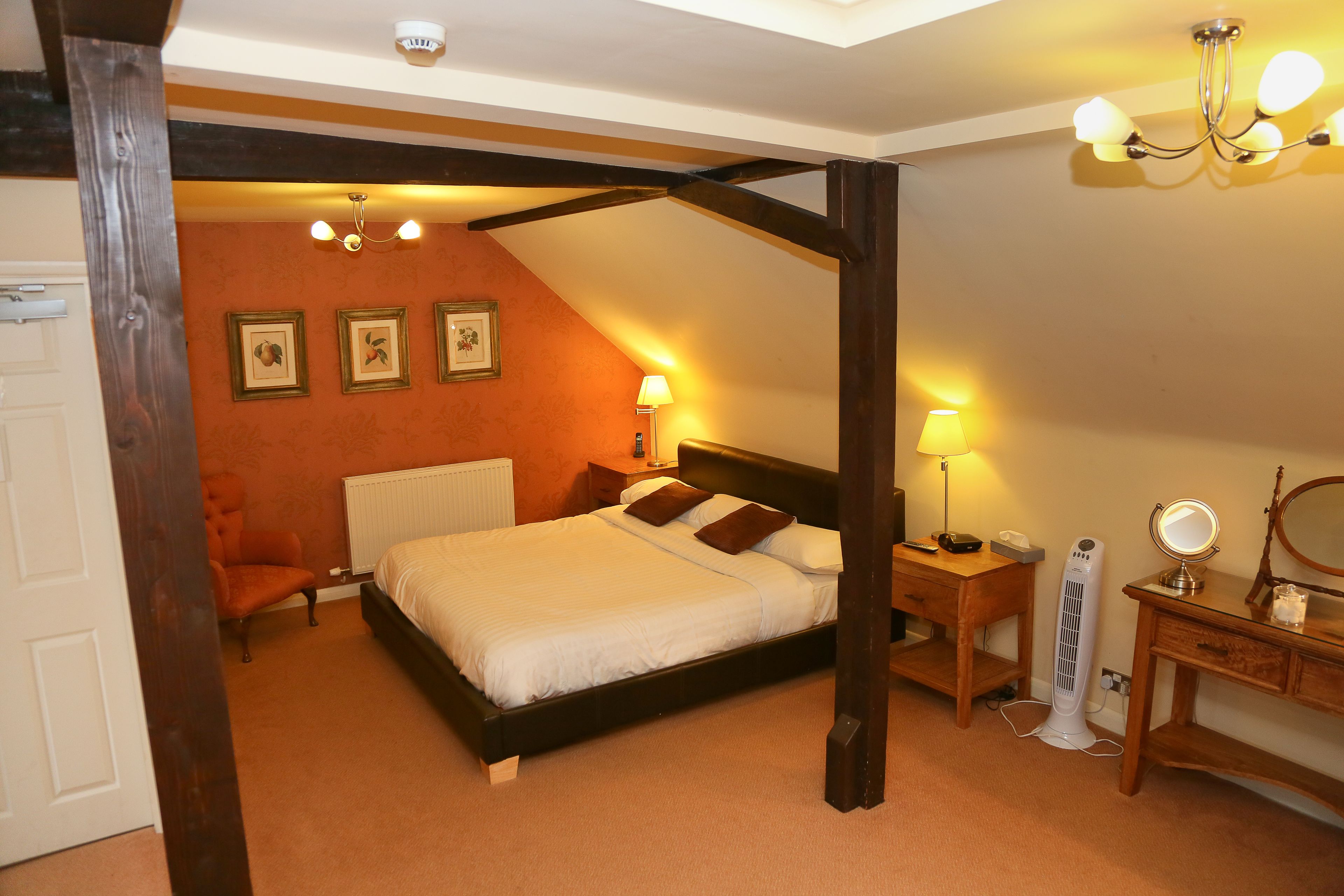 A bedroom with a four-poster bed, a nightstand with a lamp, a dresser with a mirror, and framed pictures on the wall.