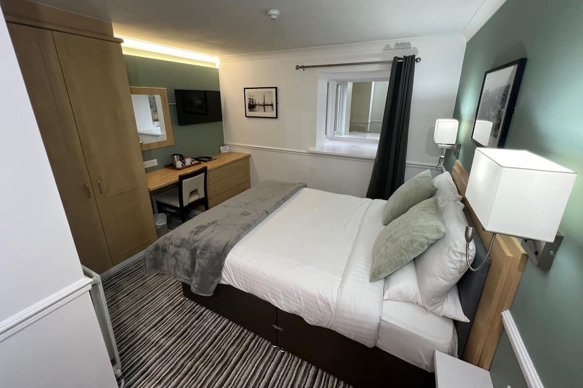 A hotel room with a bed, bedside table, lamp, and a desk with a chair.