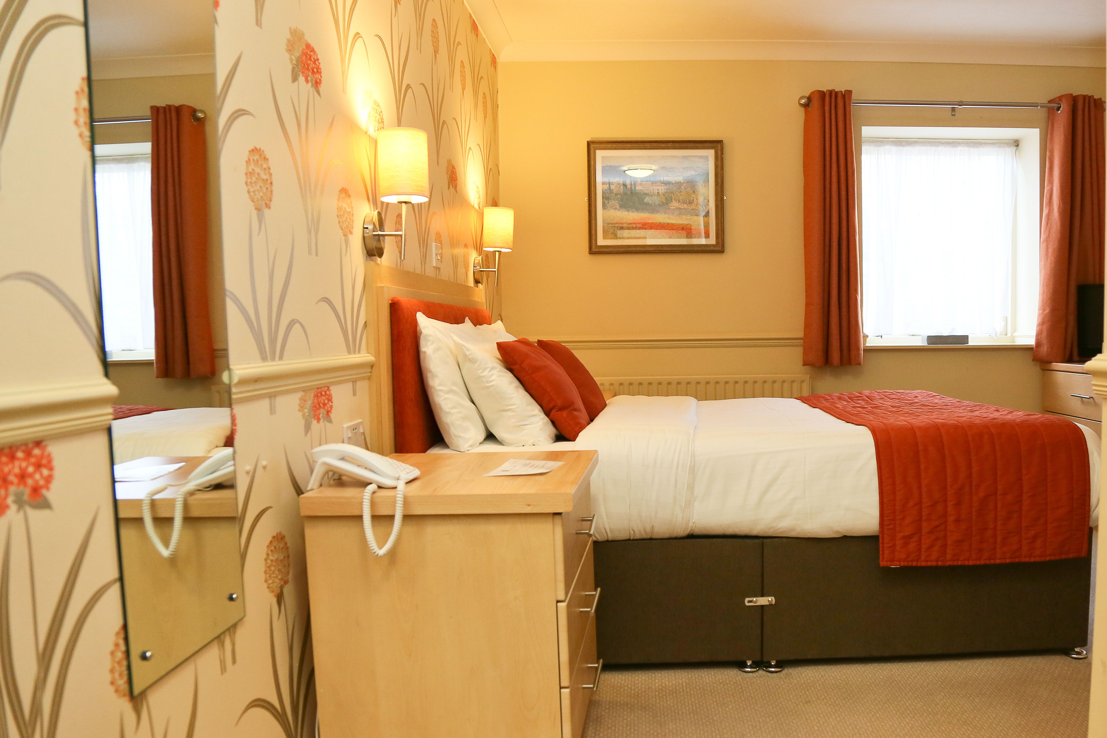 Une chambre d’hôtel avec un lit, un bureau avec un téléphone, un miroir et une fenêtre avec des rideaux.