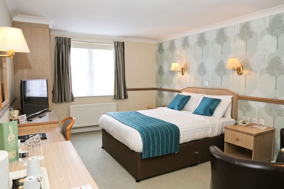A hotel room with a bed, bedside table, lamp, TV, and a desk with a chair.