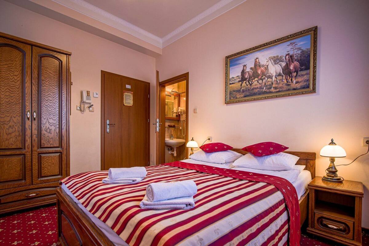Une chambre d’hôtel avec un grand lit, une armoire en bois, une salle de bain avec un miroir et un tableau représentant des chevaux accroché au mur.