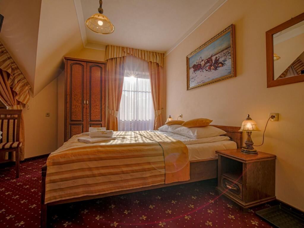A hotel room with a large bed, a wooden wardrobe, a painting on the wall, and a lamp on the nightstand.