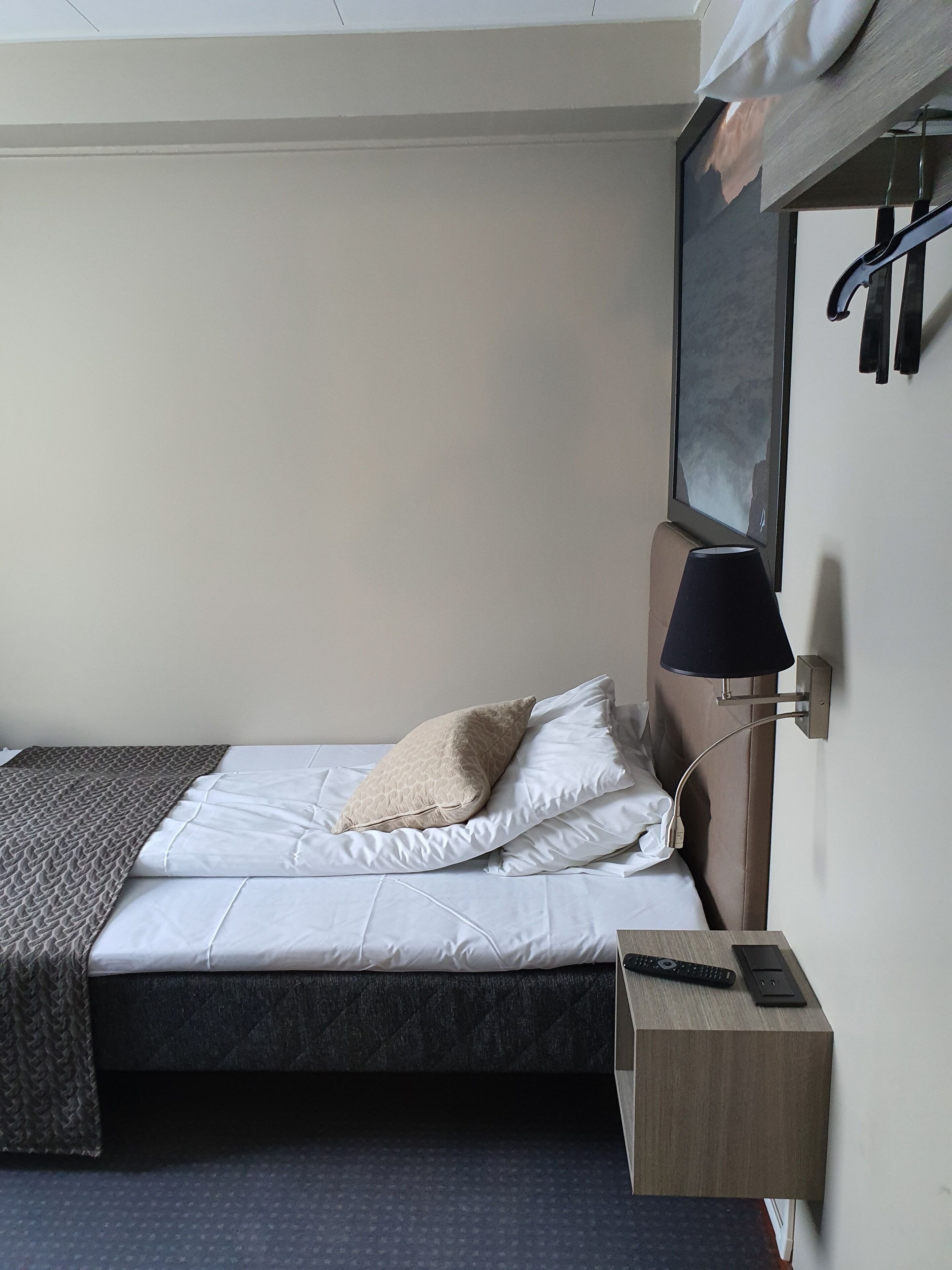 Et lille hotelværelse med en enkeltseng, et natbord med lampe, et lille trænatbord og et spejl monteret på væggen.