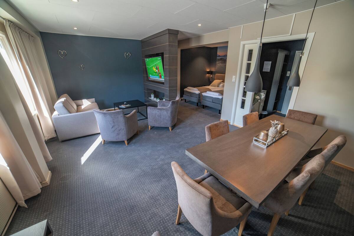 Une chambre d’hôtel moderne dotée d’une grande télévision à écran plat, d’un coin repas avec une table et des chaises en bois, d’un canapé et d’un lit avec une table de chevet.
