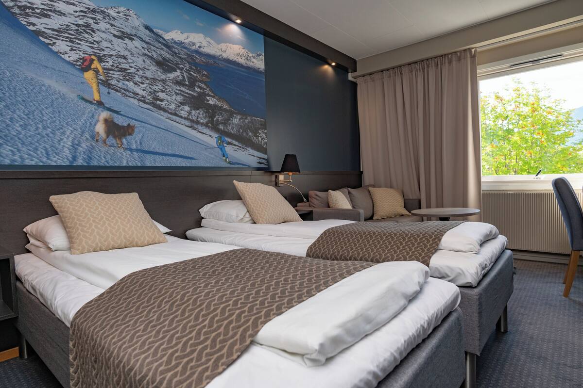 Une chambre d’hôtel avec deux lits, un grand téléviseur fixé au mur qui diffuse une image de montagnes enneigées, une petite table et une chaise.