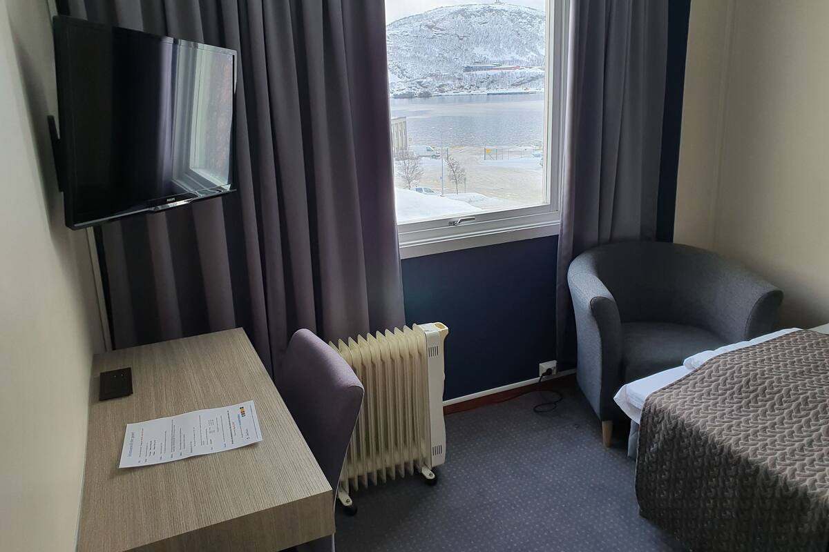 Une chambre d’hôtel avec un lit, une chaise, une table avec un menu, une télévision et une fenêtre donnant sur un paysage enneigé.