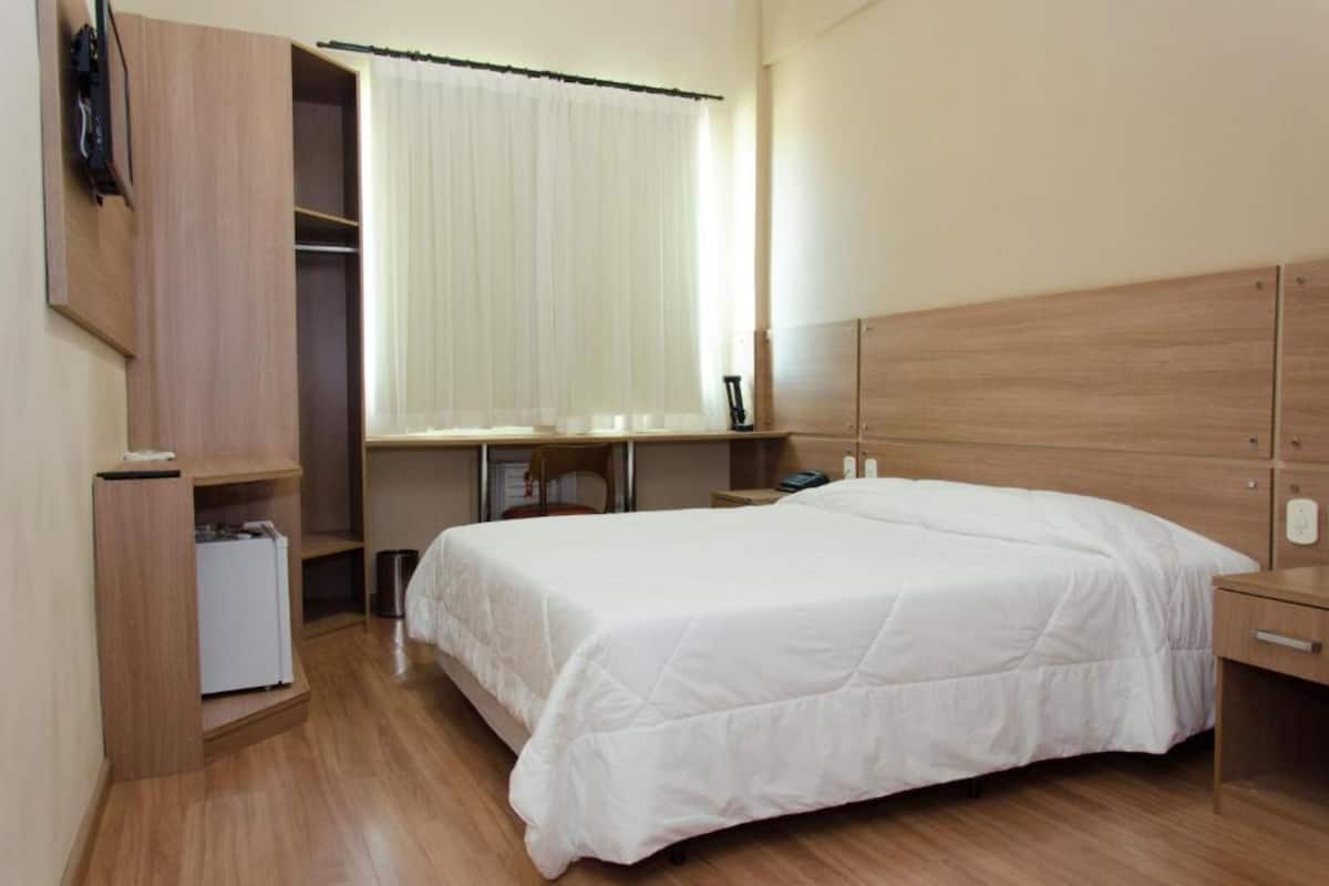 Una habitación de hotel con cama, escritorio, silla, armario y ventana con cortinas.