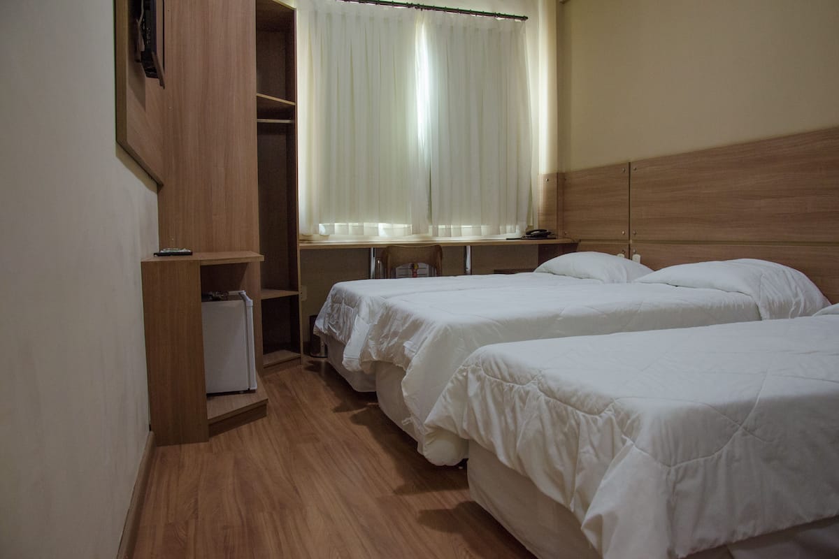 Habitación de hotel con dos camas individuales, suelo de madera, un armario y una ventana con cortinas traslúcidas.