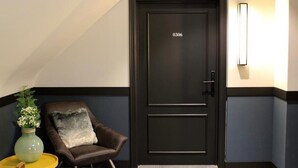 A dark-colored door with the number 0306, a brown leather chair with a grey pillow, a small yellow table with a green vase, and a wall-mounted light fixture.