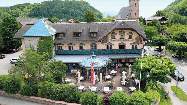 A traditional building with a tower, a covered outdoor seating area, and a view of a forested hill. - Gasthof Schorn (Groedig)