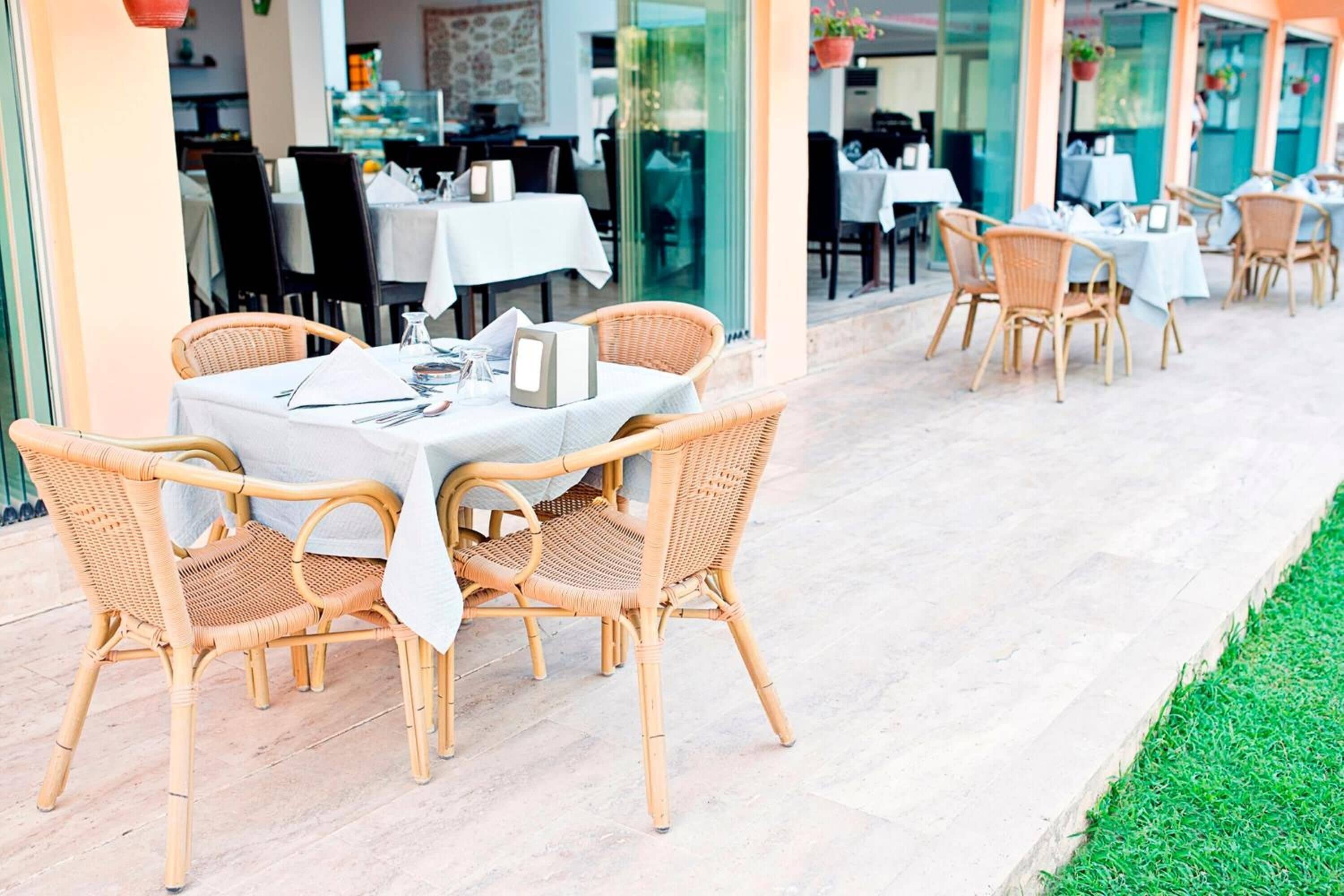 Un área de comedor al aire libre con mesas y sillas de mimbre, piso de baldosas y una pared de vidrio que separa el comedor del interior.