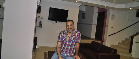 A man sitting on a brown leather sofa in a modern lobby with a flat-screen TV mounted on the wall and a staircase in the background.