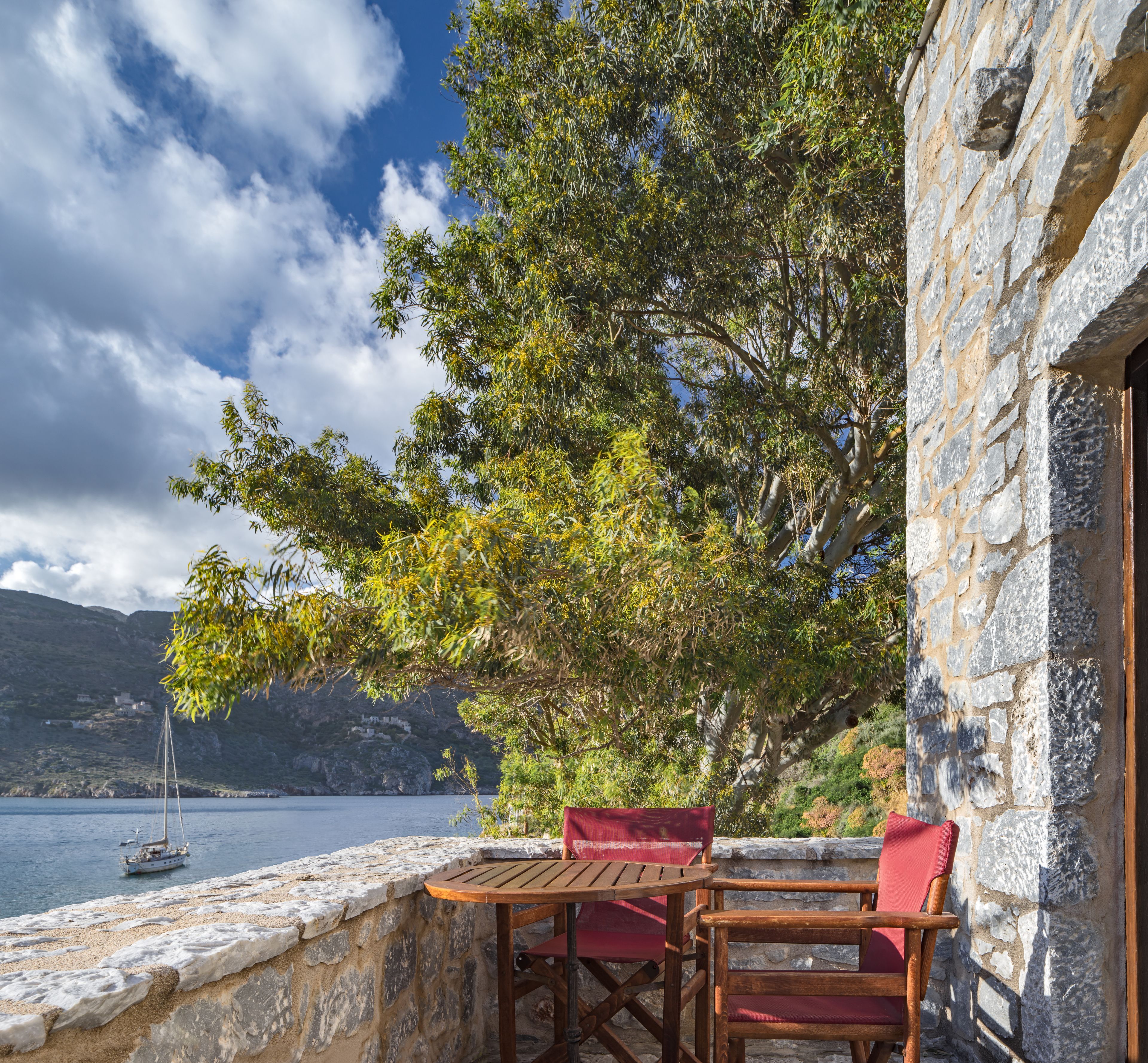 A stone building with a wooden table and chairs by a stone wall overlooking a body of water with a sailboat.