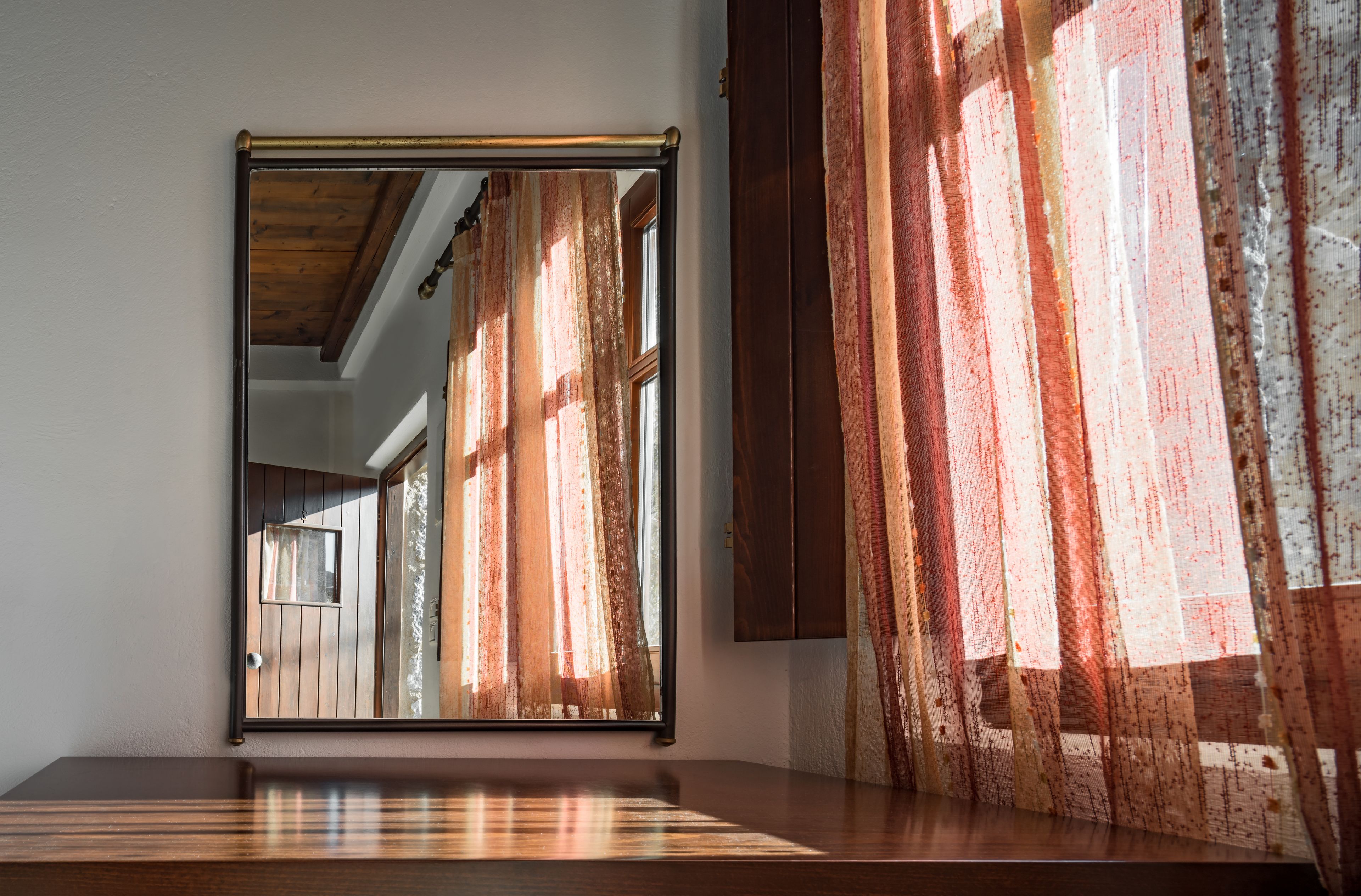 A mirror with a wooden frame, reflecting a room with a wooden ceiling and a window with curtains.