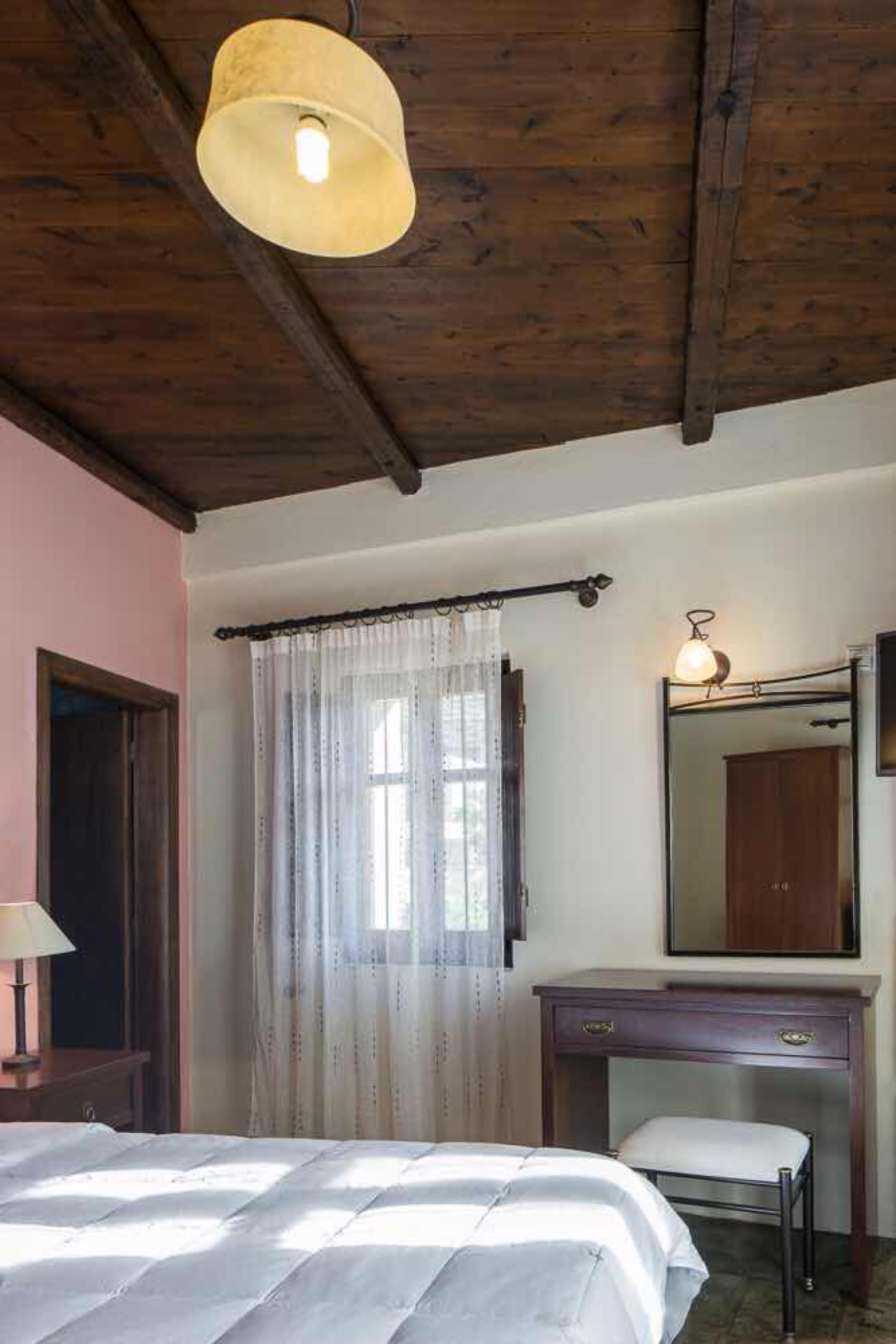 A bedroom with a bed, a desk, a chair, a mirror, and a hanging light fixture.