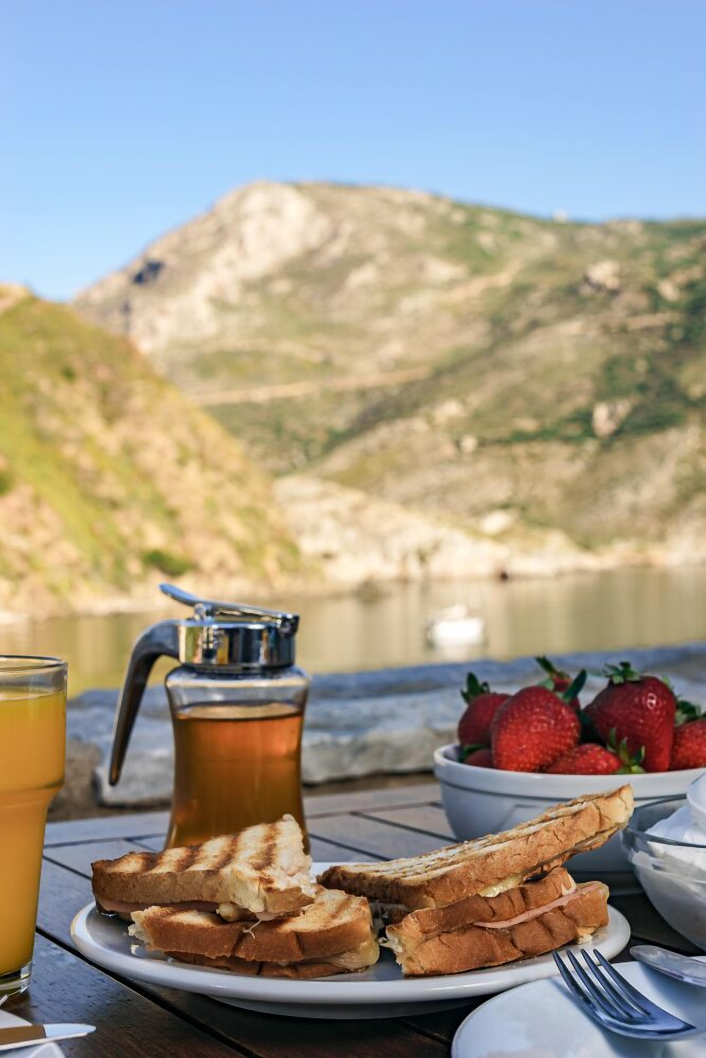 Frühstückssituation mit Toast, Erdbeeren, einer Kanne und einem Glas Saft auf einem Tisch mit Blick auf Berge im Hintergrund.