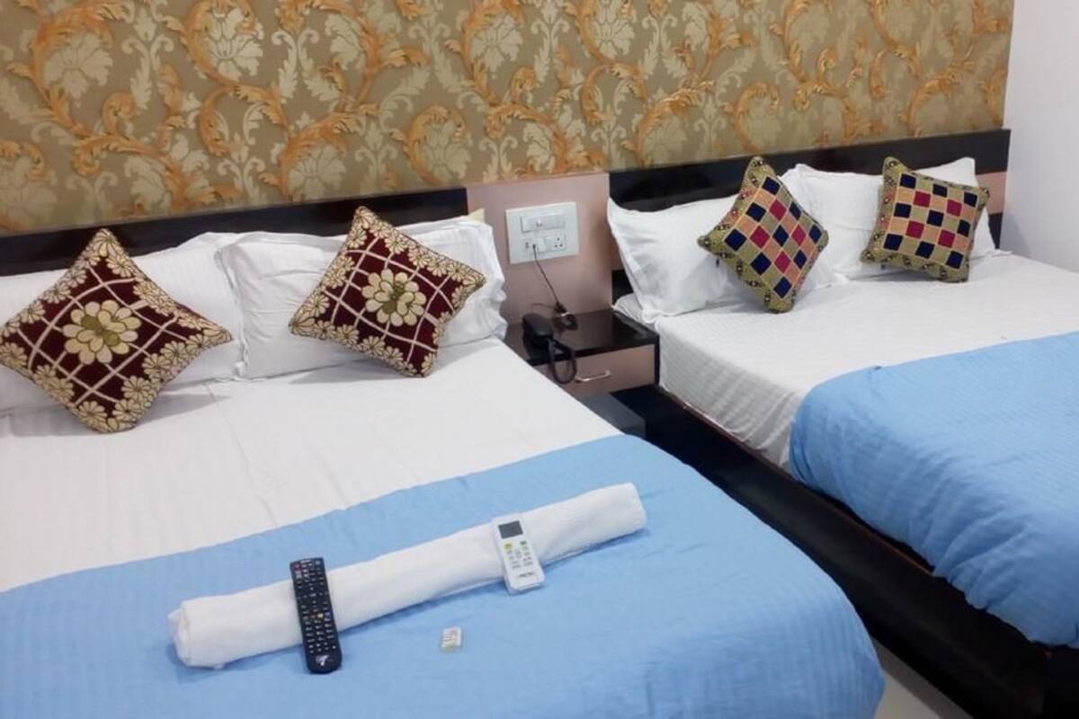 A hotel room with two beds, each with a blue bedspread and white linens. There are decorative pillows on the beds, a bedside table with a lamp, and a remote control on the bed.