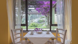 A dining table with chairs, a window with purple flowers, and a view of greenery and hills. - Argentiera Inn (Sassari)