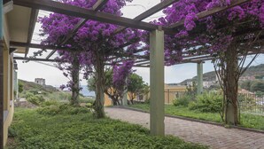 A covered garden area with purple flowers, a brick pathway, and a view of buildings in the background. - Argentiera Inn (Sassari)