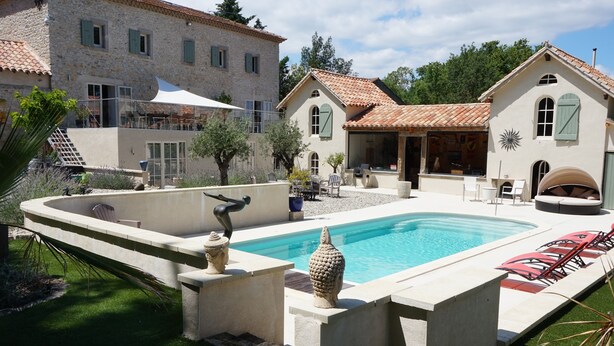 Une piscine bordée de pierres, un espace extérieur aménagé avec des sièges et un bâtiment doté d’un toit en tuiles.