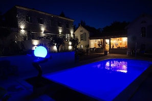 Una piscina con iluminación azul, un edificio de piedra con ventanas iluminadas y una zona de bar con señalización visible.