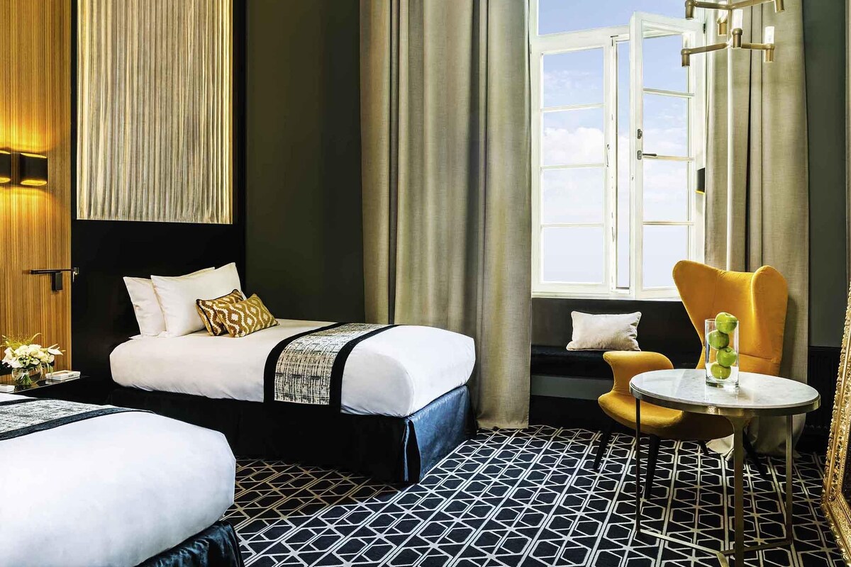 A hotel room with a bed, a chair, a small table, and a window with curtains.