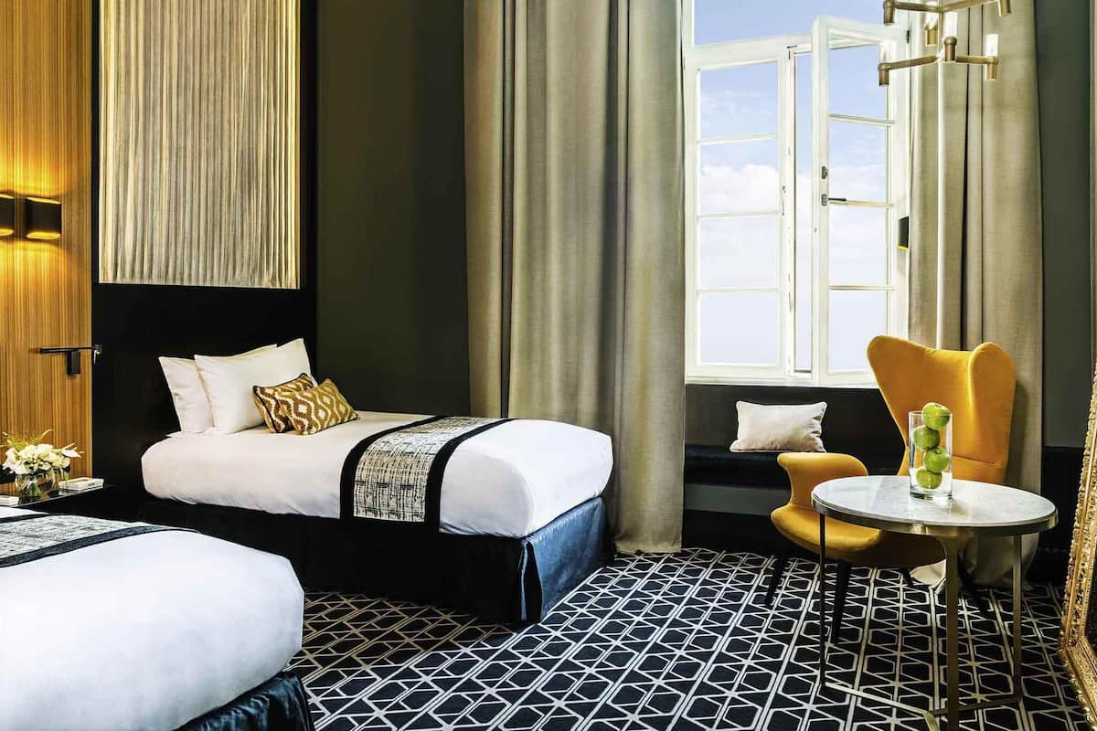 Habitación de hotel con cama, una silla, una mesita y un ventanal con cortinas.
