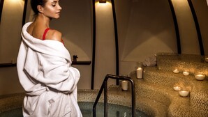 Una persona en una bata blanca, de pie junto a una piscina de spa con un área de asientos texturizada y velas.