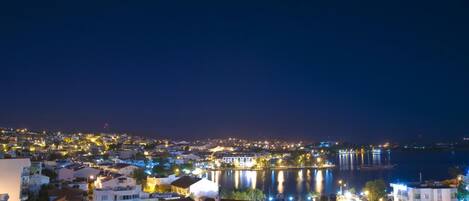 A nighttime view of a coastal town with buildings along the waterfront, a bridge, and a calm body of water reflecting city lights.