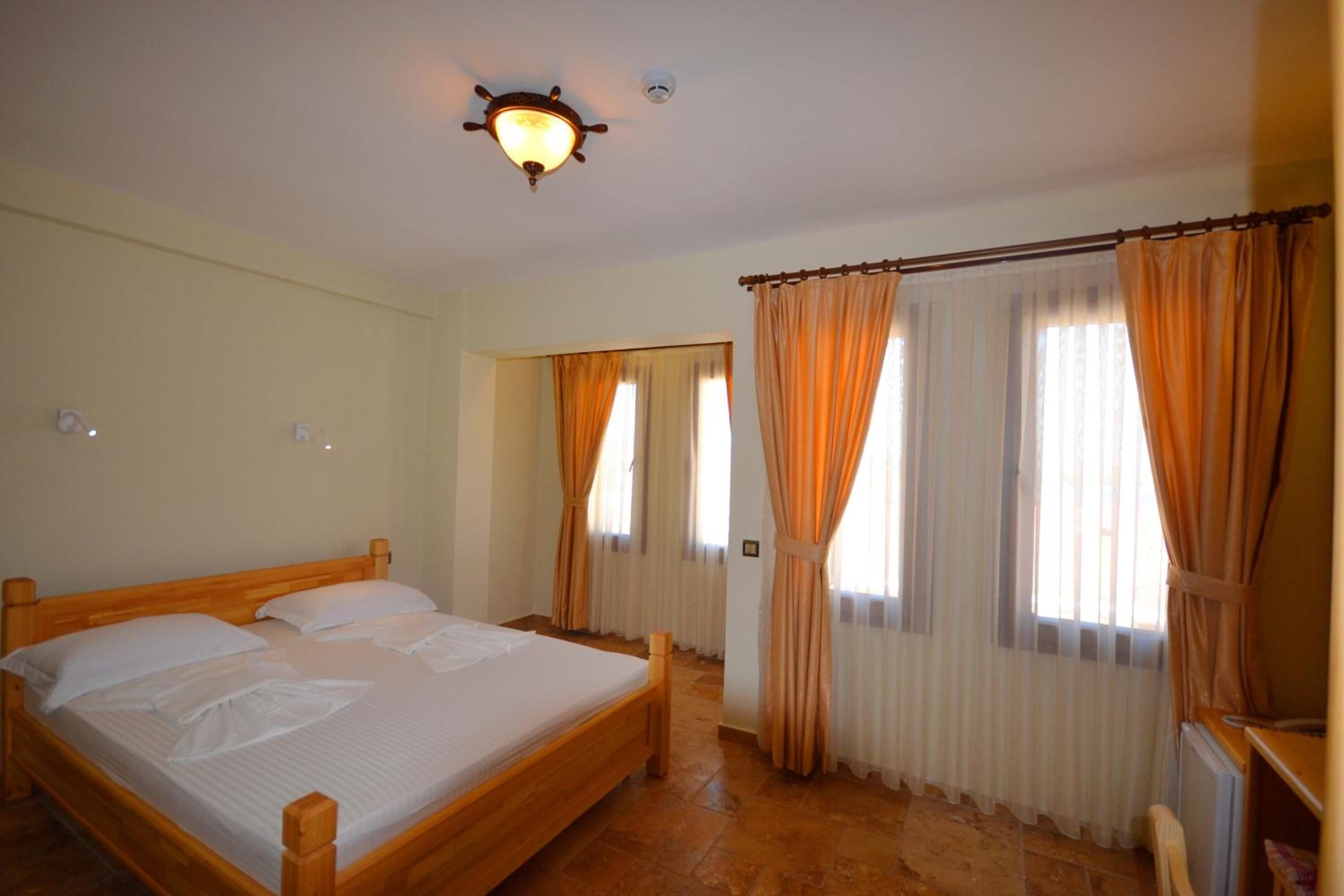 A bedroom with a wooden bed, two windows with curtains, a ceiling light fixture, and a tiled floor.