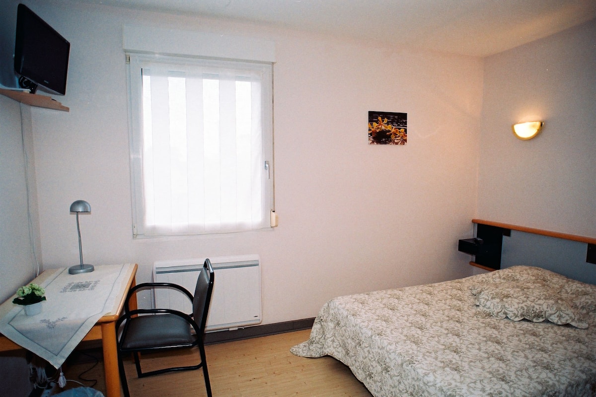 Un dormitorio con cama, escritorio y silla, radiador, ventana con persianas, e iluminación fijada a la pared.