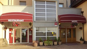 Un edificio que alberga un restaurante y un hotel. El restaurante tiene un toldo rojo y una cesta de flores colgando sobre la entrada. El hotel también tiene un toldo rojo y una cesta de flores colgando sobre la entrada.