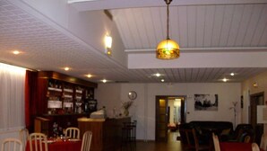 Un comedor con mesas dispuestas para dos, una zona de bar con estantes llenos de botellas, y una sala de estar con un sofá y sillones.