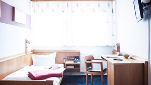 A hotel room with a bed, a desk, a chair, a desk lamp, a clock, and a window with curtains.