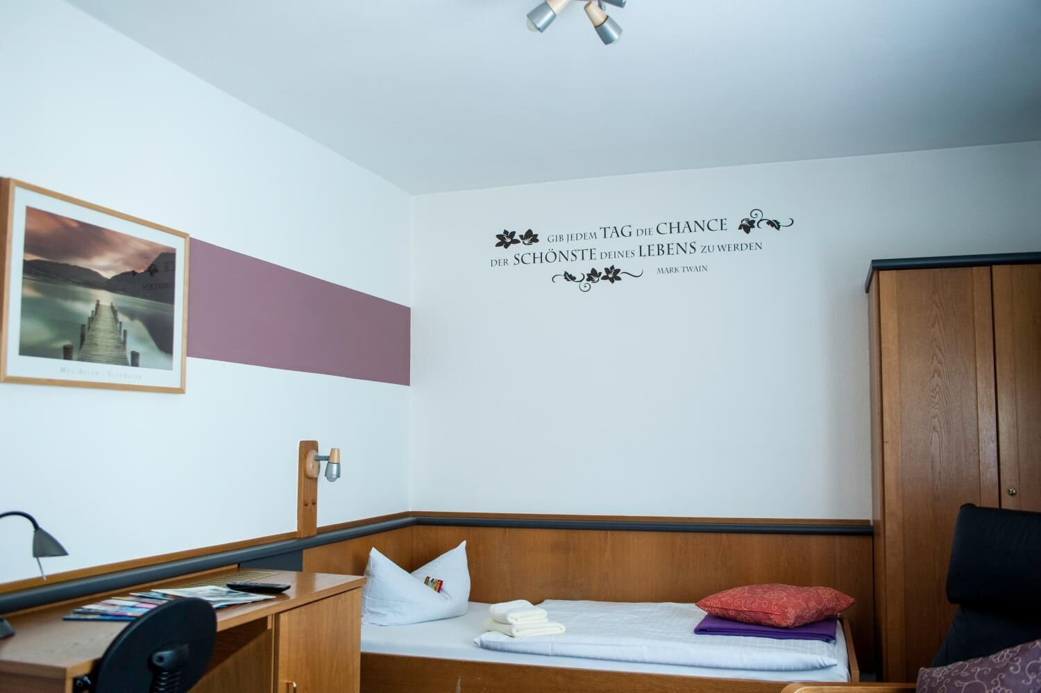 A hotel room with a bed, a desk, a chair, a wardrobe, and a wall with a quote.