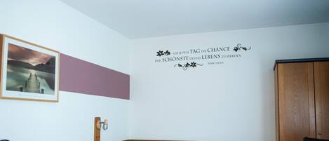 A hotel room with a bed, a desk, a chair, a wardrobe, and a wall with a quote.
