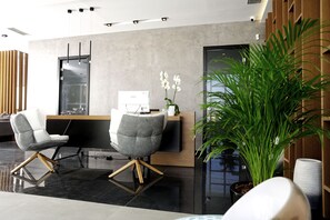 A modern reception area with two grey chairs, a wooden desk, a computer, and a large green plant.