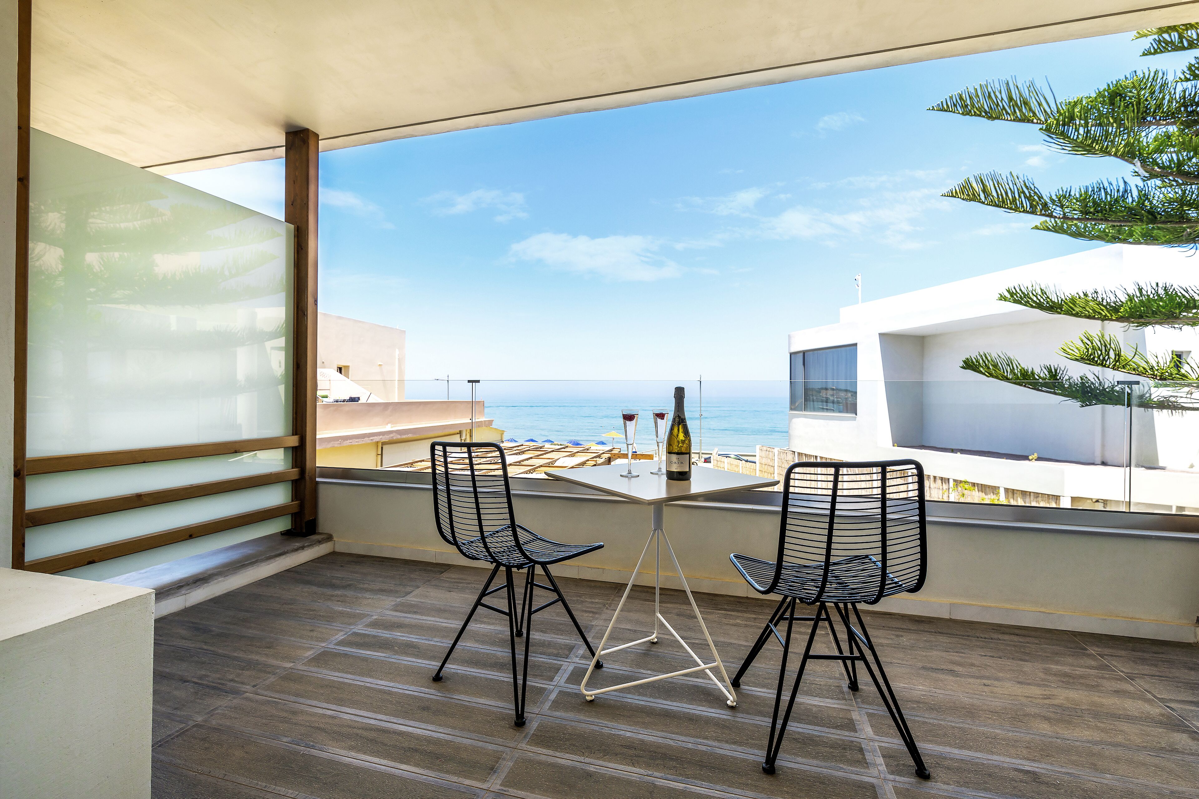 A balcony with two chairs, a table, and a bottle of wine, overlooking a coastal view.