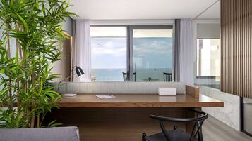 Habitación moderna con escritorio de madera, una silla, un ventanal con vistas al mar y una planta en maceta.