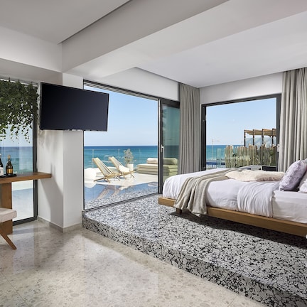 A modern bedroom with a large bed, a desk, and a TV. It features a balcony with a view of the sea.