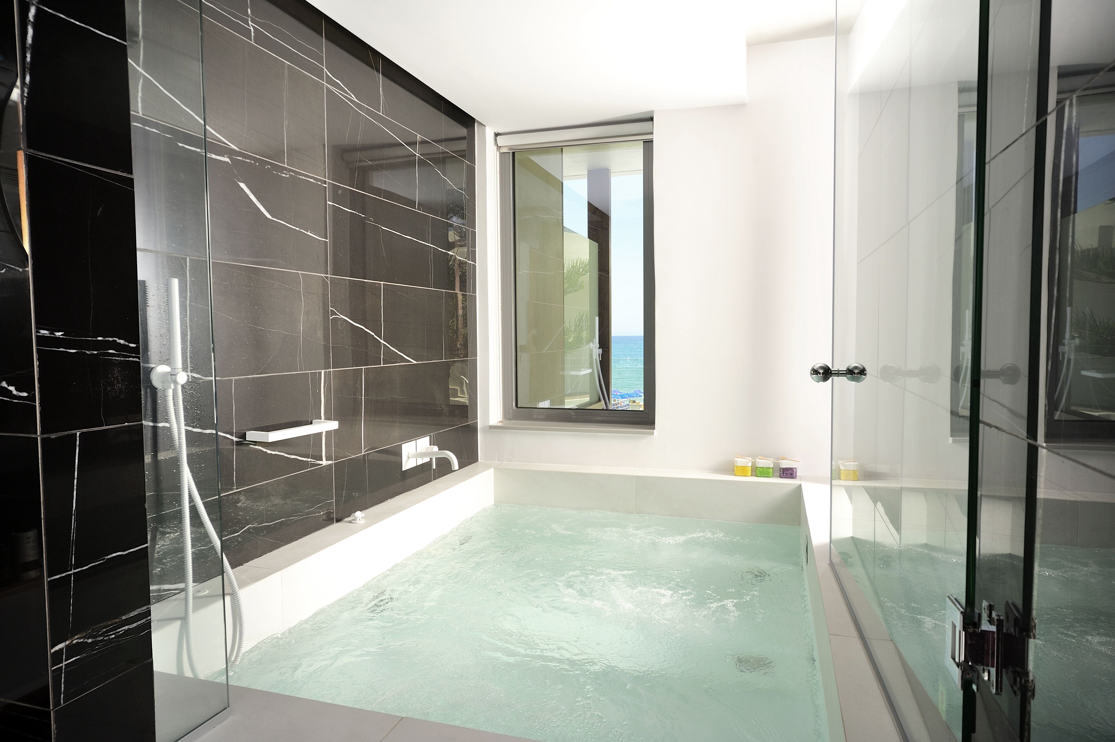 A modern bathroom with a glass-enclosed shower, a bathtub with a view, and a window overlooking the sea.