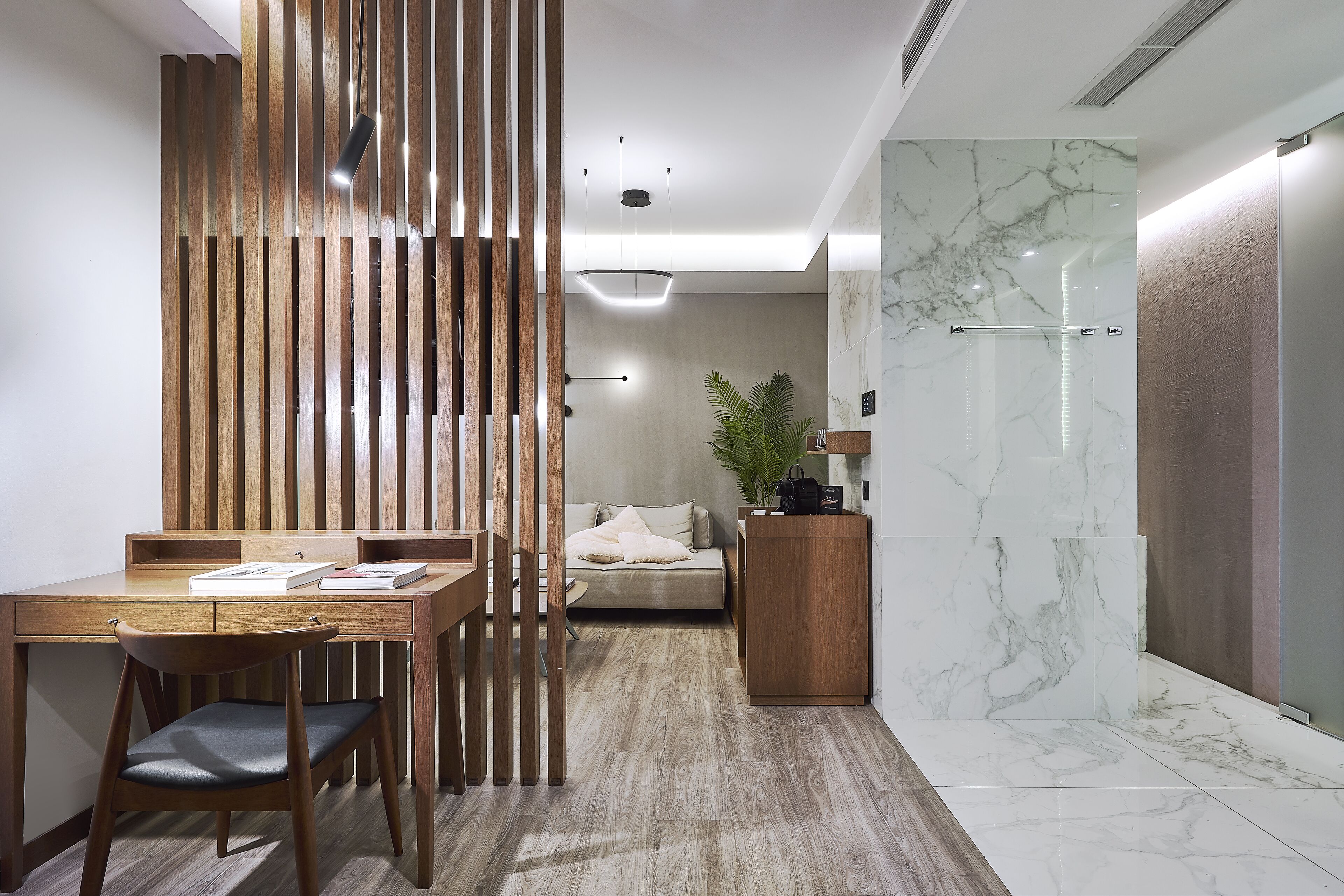 Un interior moderno con un escritorio de madera, una silla acolchada, un sofá, una planta y una zona de ducha con azulejos de mármol.