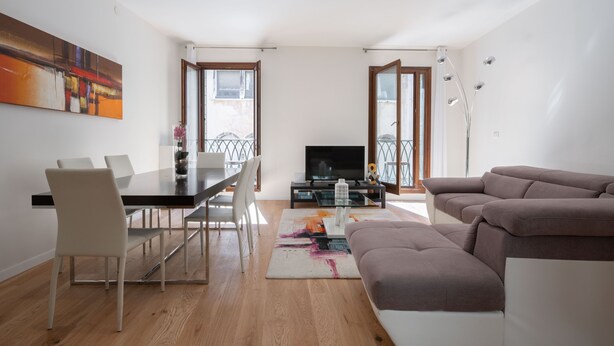 A modern living room with a dining area, a sofa, a TV, and a balcony with a railing. - San Marco Prestige with Terrace by Wonderful Italy (Venice)
