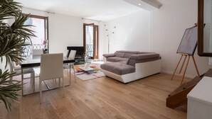 A modern living room with a sofa, a dining table with chairs, a painting on easels, and a large window. - San Marco Prestige with Terrace by Wonderful Italy (Venice)
