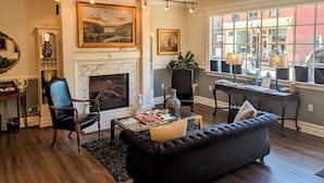 A living room with a fireplace, a sofa, a coffee table, and a view of the street through large windows.